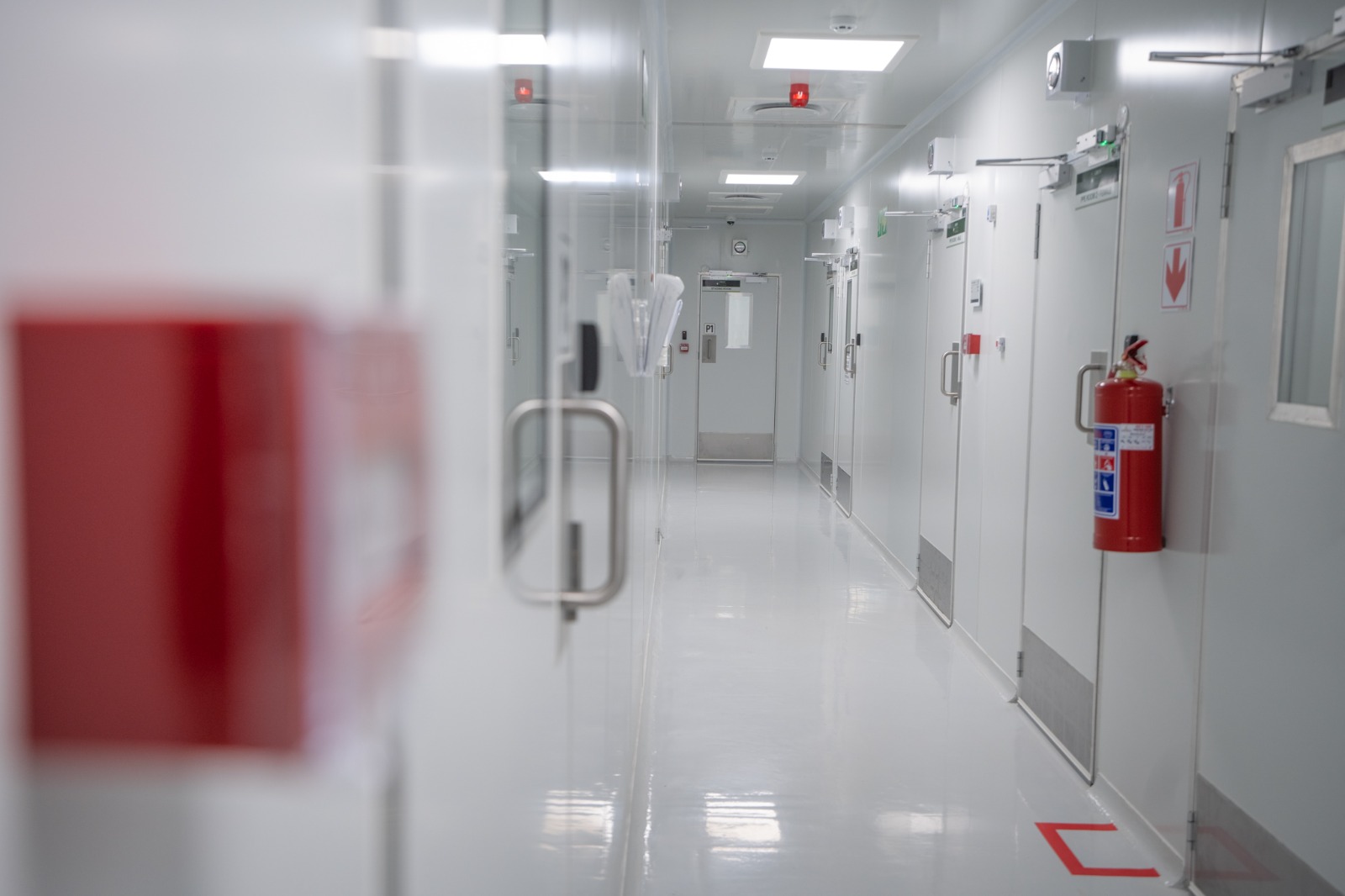 A classified white corridor lit from above, cleanroom doors and a fire extinguisher along the right-hand wall, looking towards the door marked P1