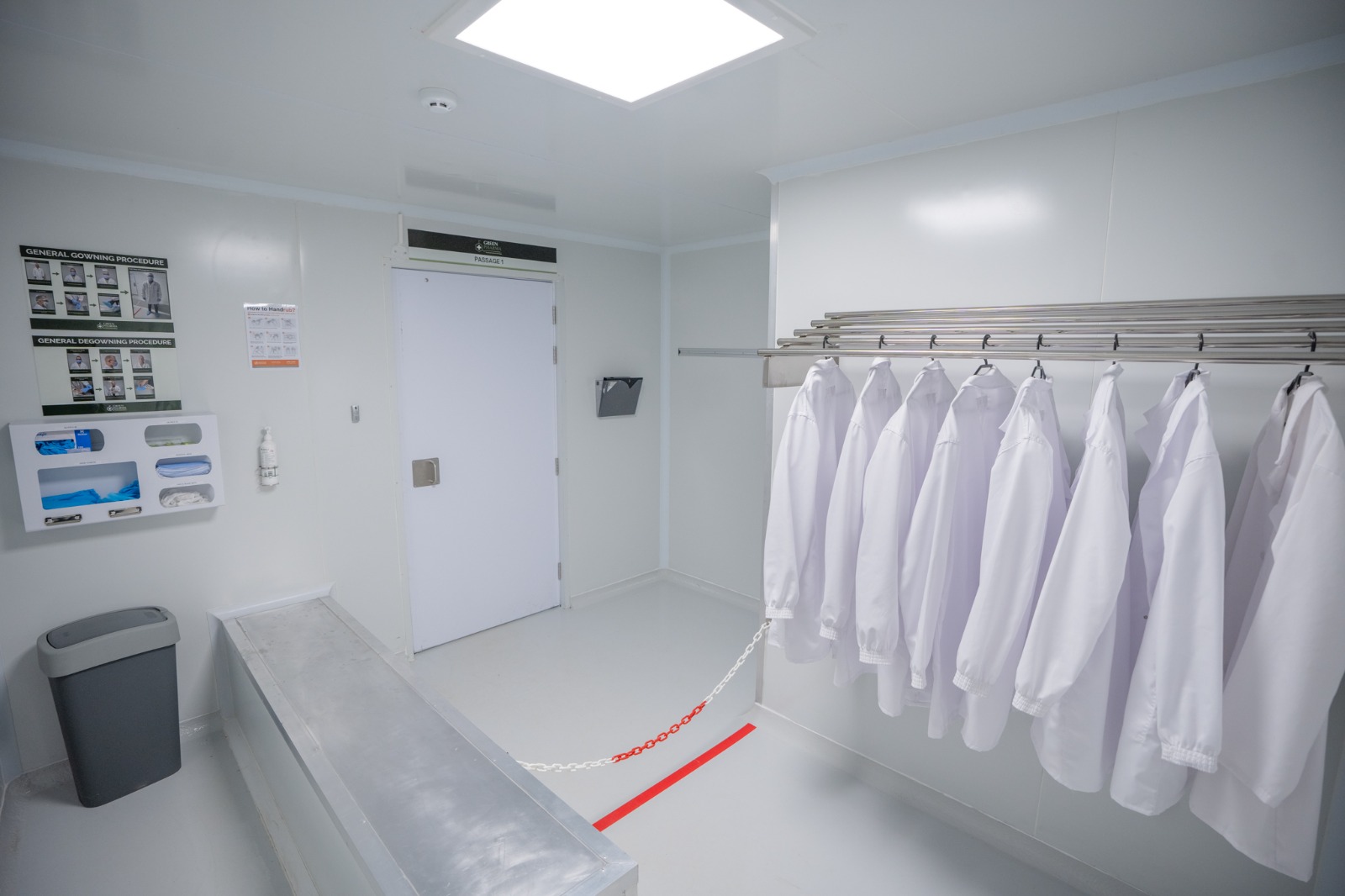 A gowning room with clean white coats hanging on a rail, a step-over bench, gowning and de-gowning procedure posters on the wall, and a red chain marking the clean side