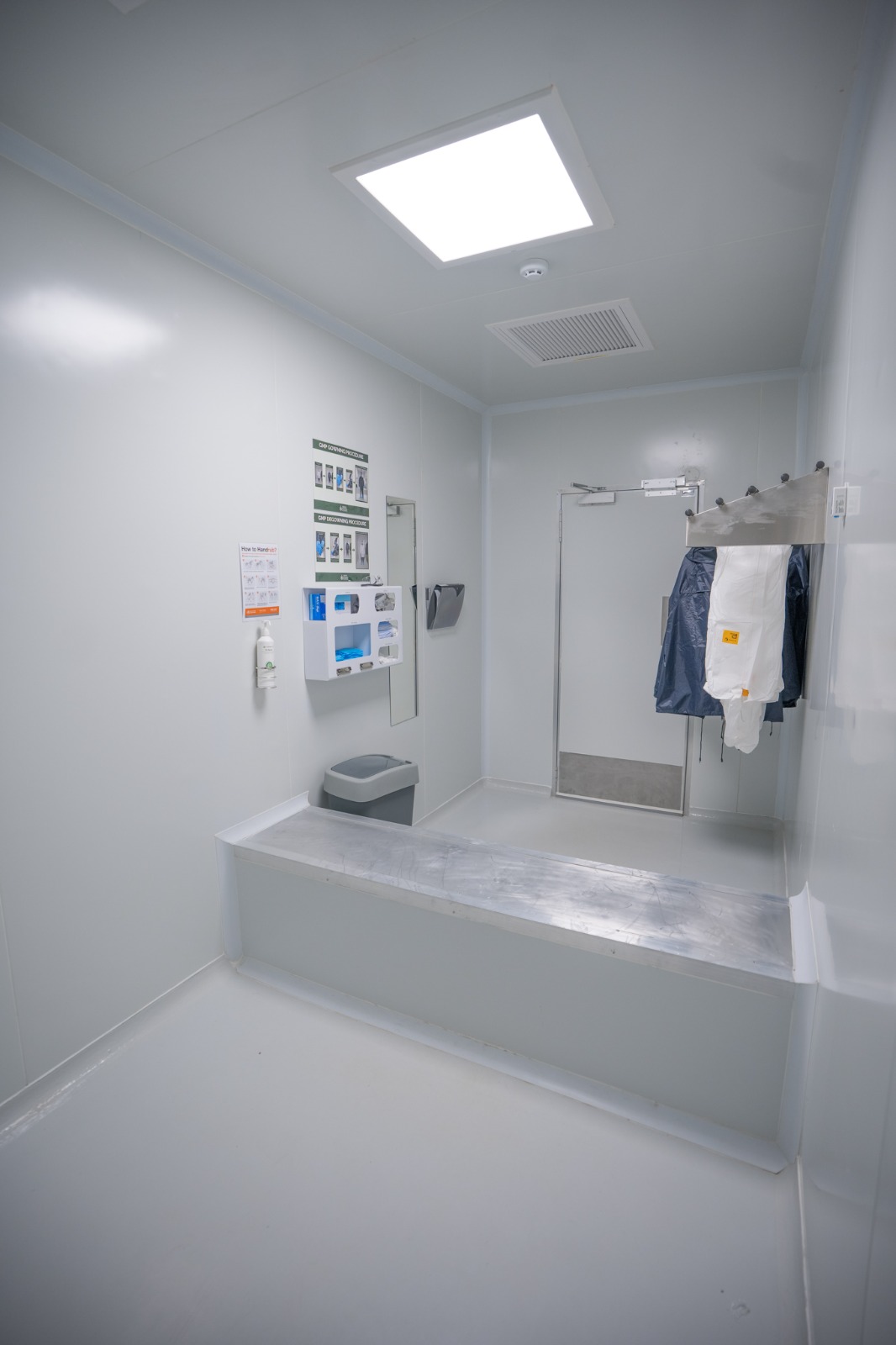 A gowning room from the doorway, step-over bench and mirror, hand hygiene station and the printed gowning procedure on the wall