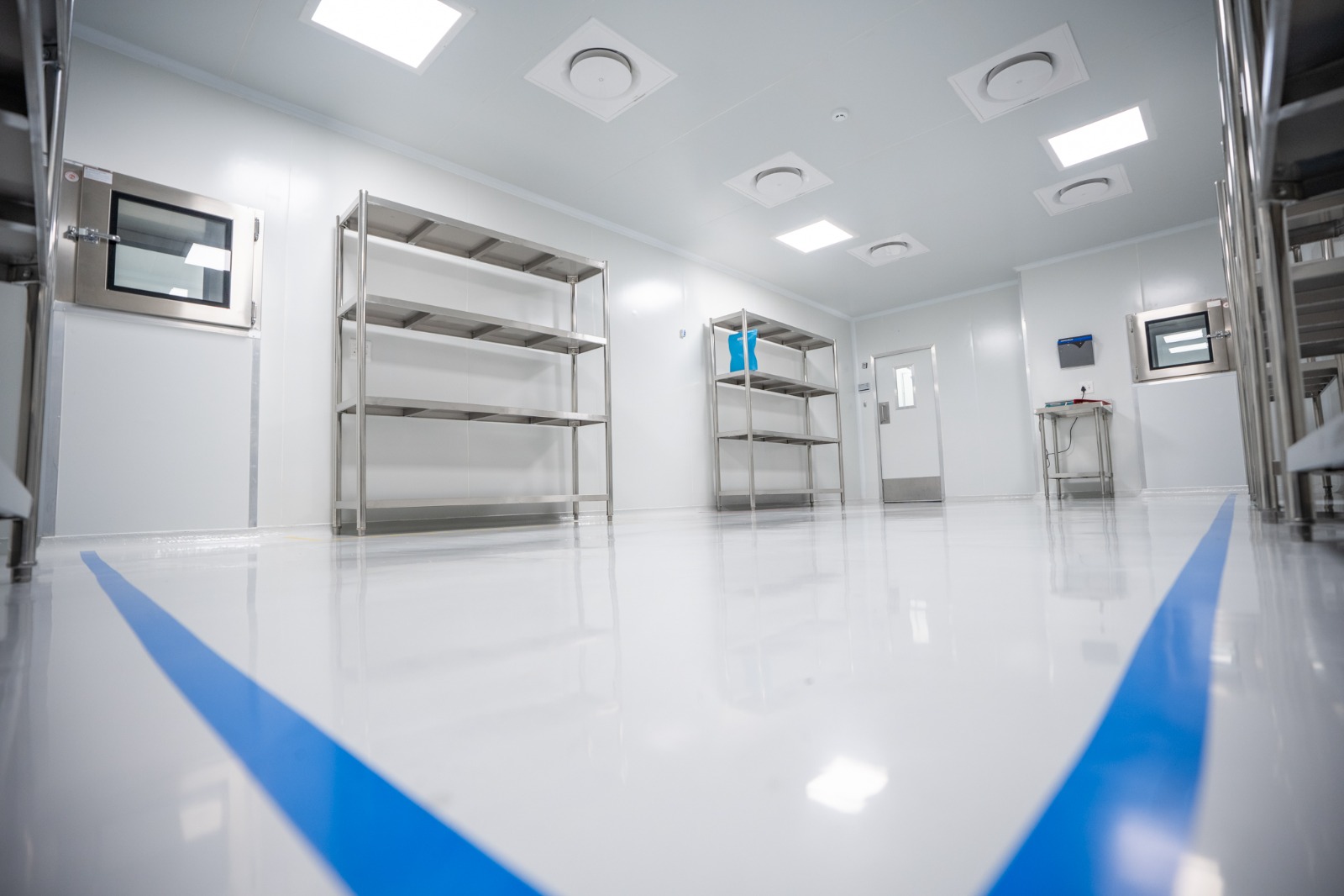 A staging room seen from floor level, empty stainless racking against the wall and two blue demarcation lines running away across a mirror-finish floor