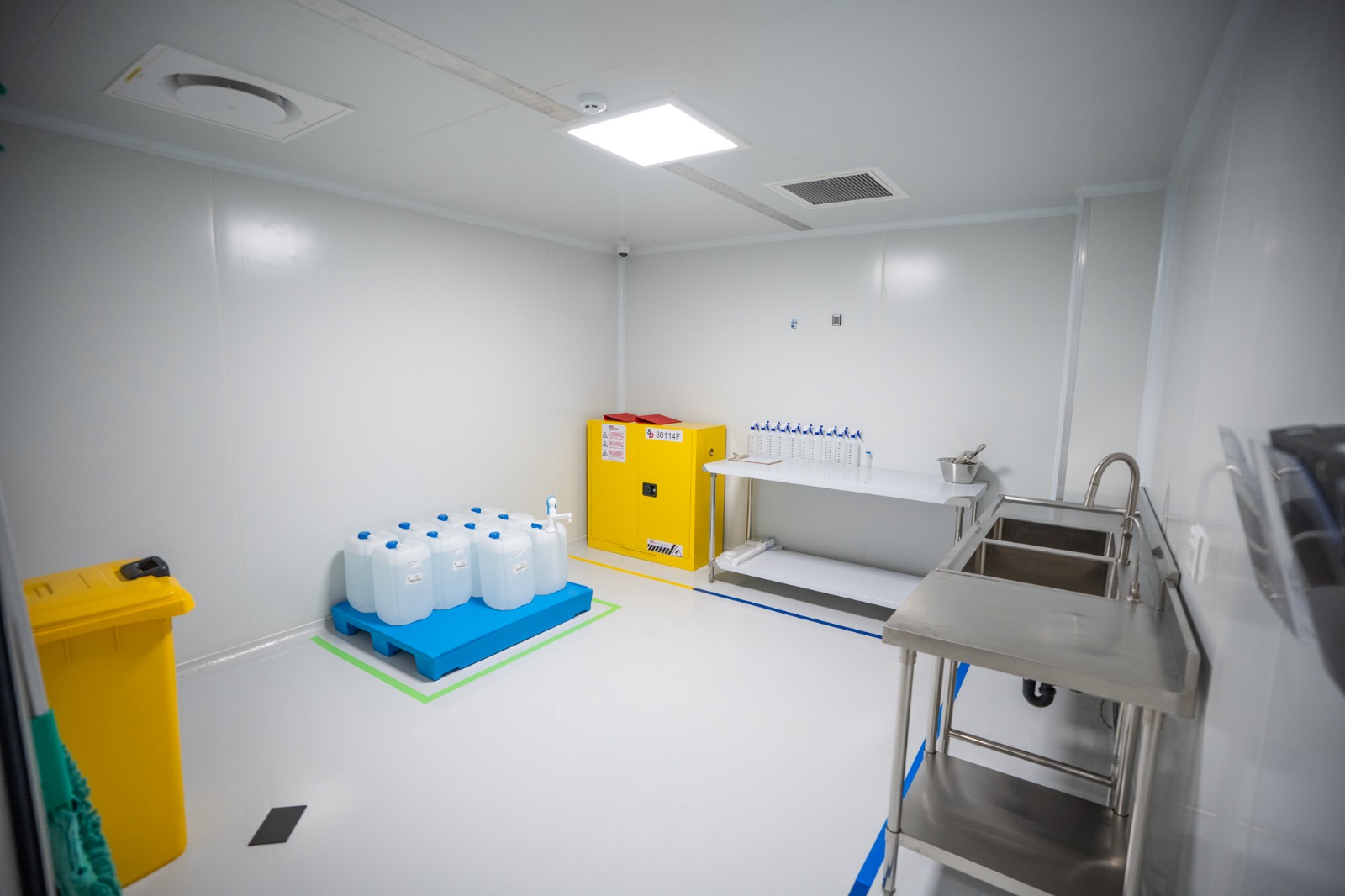 The washbay: a yellow flammables cabinet, sanitiser bottles ranked on a shelf, drums on a spill pallet inside green demarcation, and a stainless double sink